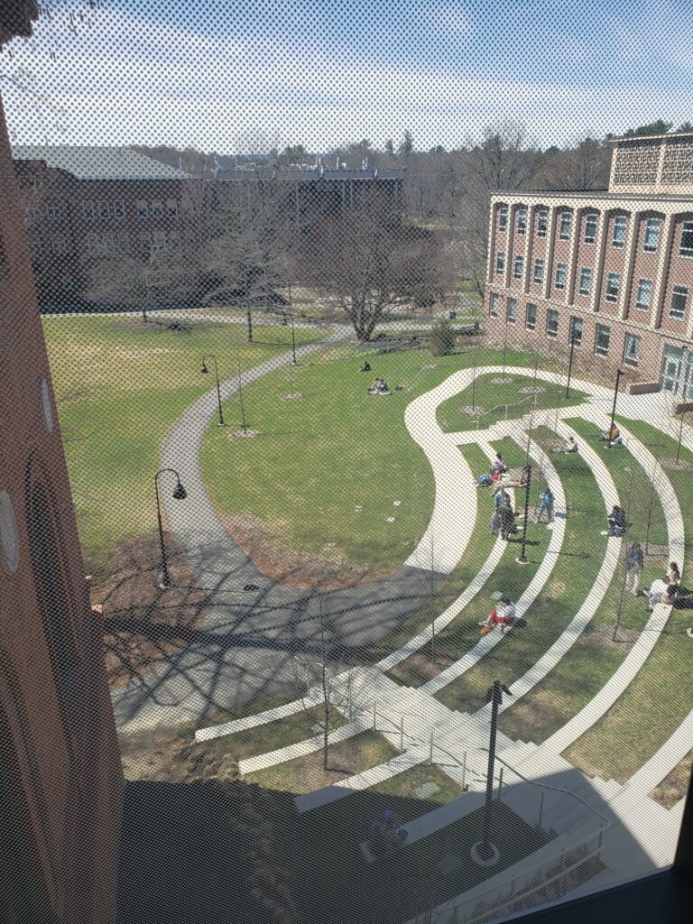 Outside of the screen on the third floor of the Nielson Library, outlooking a quad space of Smith College campus. Students are scattered around a multi-tiered amphitheater space.